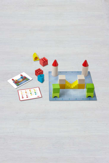 Eine Tischplatte mit einem Bauklotzspiel für Kinder, mit einer blauen Gitterbasis, bunten Holzklötzen, die zu Türmen und Bögen zusammengesetzt werden, losen Klötzen und zwei Anleitungskarten mit Bildern und Diagrammen.