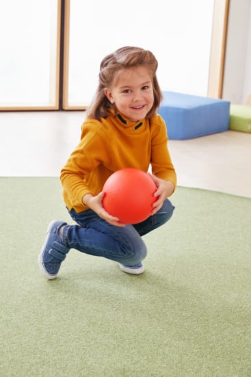 Ein junges Mädchen mit hellbraunem Haar, das einen senfgelben Pullover und Jeans trägt, hockt auf einem grünen Teppich und hält einen roten Ball in der Hand. Sie lächelt und blickt leicht nach oben.