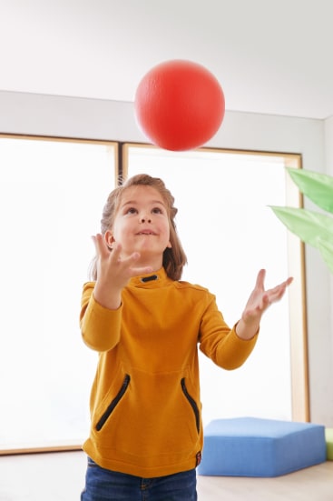 Ein Kind in einem gelben Pullover wirft einen großen roten Ball in die Luft und steht vor großen Fenstern und einer Grünpflanze.