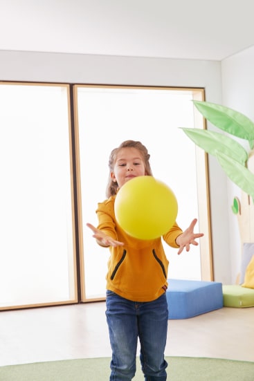 Ein kleines Kind in einem gelben Kapuzenpulli und Jeans fängt oder wirft einen großen gelben Ball in einem Haus. Im Hintergrund sind große Fenster und ein Teil einer Pflanzendekoration zu sehen.