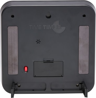 Die Rückansicht eines schwarzen, quadratischen Geräts mit der Aufschrift TIME TIMER, das ein Batteriefach, einen roten Schalter, zwei vertikale Gummistreifen und zwei ausklappbare Ständer an den unteren Ecken aufweist.