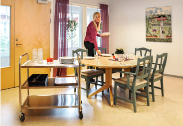 Eine Frau steht an einem runden Esstisch und deckt Teller ein. In der Nähe steht ein Servierwagen aus Metall mit Gläsern, Tabletts und Tellern. Der Raum hat Holzstühle, eine Tür, Fenster mit Vorhängen und ein Gemälde an der Wand.