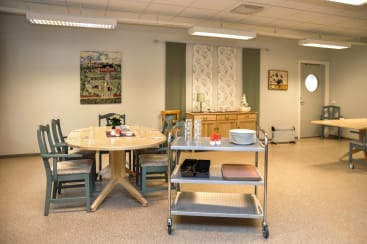 Ein Essbereich mit einem runden Holztisch mit Tellern und Gläsern, grünen Stühlen, einem Servierwagen aus Metall mit Geschirr und hellen Schränken an der Wand, über denen dekorative Kunst hängt.