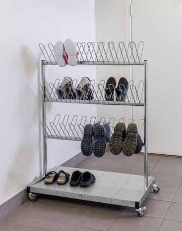 Ein Schuhregal aus Metall mit drei Fächern enthält verschiedene Paar Schuhe, darunter Sandalen, Turnschuhe und Stiefel. Das Regal steht an einer weißen Wand auf einem gefliesten Boden in der Nähe einer geschlossenen Tür.