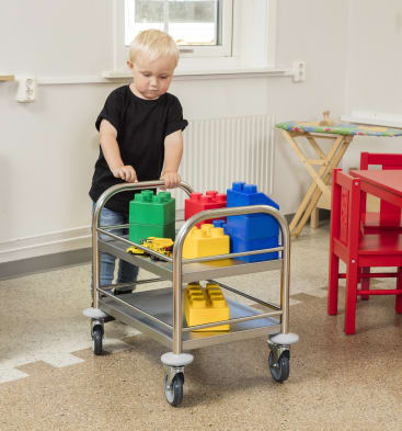 Ein kleines Kind schiebt einen Metallwagen mit großen, bunten Plastikbausteinen und einem Spielzeuglaster in einem Klassenzimmer mit roten Möbeln und einem Fenster im Hintergrund.