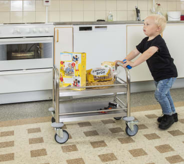 Ein kleines Kind schiebt einen Metallwagen mit Lebensmitteln wie Müsli, Brot und einem Korb in einer Küche. Der Wagen hat drei Fächer und große Räder. Die Küche ist mit einem Herd und weißen Schränken im Hintergrund ausgestattet.