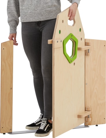 Eine Person, die einen grauen Pullover, dunkle Jeans und schwarze Turnschuhe trägt, steht neben einer teilweise montierten Holzkonstruktion mit einem grünen sechseckigen Ausschnitt und zwei aufklappbaren Platten.
