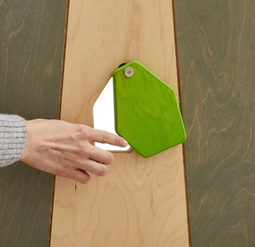 Eine Person, die einen grauen Pullover trägt, dreht eine grüne, fünfeckige Abdeckung, die auf einer hölzernen Oberfläche angebracht ist, und enthüllt einen kleinen rechteckigen Spiegel darunter.