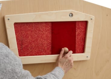 Die Hand einer Person hält einen Holzknauf, der an einer Schiebetafel in einem hellen Holzrahmen befestigt ist. Das Paneel ist rot mit feinen Linienmustern, und der Hintergrund ist ebenfalls aus hellem Holz gefertigt.