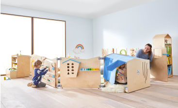 Eine Frau und zwei Kinder spielen in einem hellen Raum mit Spielgeräten aus Holz. Ein Kind hockt an einem Spielzeugtor, das andere und die Frau befinden sich teilweise in einer mit einem blauen Tuch ausgelegten Festung. Regale und Spielzeug sind sichtbar.