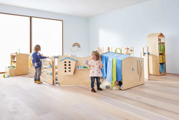 Zwei kleine Kinder spielen in einem hellen Raum mit hölzernen Spielgeräten in Form von Häusern. Spielzeug und Bücher stehen in Regalen, und ein großes Fenster lässt natürliches Licht herein. Der Boden ist aus hellem Holz.