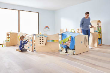 Zwei Kinder spielen mit hölzernen Spielgeräten in einem hellen, geräumigen Raum, während ein Erwachsener lächelnd daneben steht. Der Raum umfasst Regale mit Spielzeug, Bücher und einen dekorativen Regenbogen an der Wand.