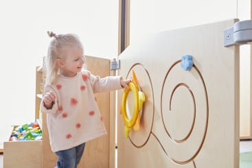 Ein kleines Kind mit blondem Haar berührt ein rundes gelbes Spielzeug, das auf einem hellen Holzspielgerät mit einem Labyrinthmuster angebracht ist. Im Hintergrund ist ein Behälter mit buntem Spielzeug zu sehen.
