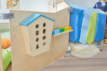 Eine hölzerne Spieltafel mit einem Haus, das Formen sortiert, und bunten Blöcken steht auf einem hellen Holzboden, neben blauen und grünen Stoffbahnen. In der Nähe liegt ein flauschiger weißer Teppich, und auf der rechten Seite ist ein Teil eines Kindes zu sehen.