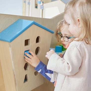 Zwei kleine Kinder setzen blaue geometrische Blöcke in die passenden Löcher auf einem hölzernen Spielbrett in Form eines Hauses. Die Kinder sind konzentriert und stehen dicht beieinander.