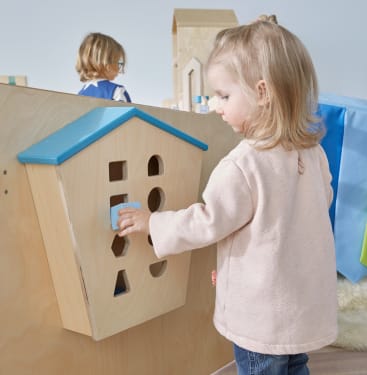Ein kleines Kind legt einen blauen Klotz in ein an einer hölzernen Trennwand befestigtes Haus zum Sortieren von Formen, während im Hintergrund ein anderes Kind zu sehen ist.