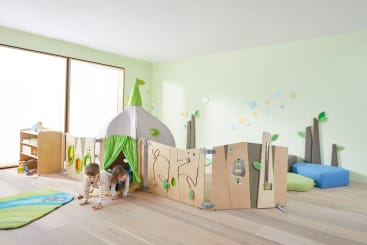 In einem hellen Spielzimmer mit hellgrünen Wänden, Holzböden und einfachen Möbeln krabbeln zwei kleine Kinder aus einer kleinen zeltartigen Spielstruktur, die von einem hölzernen Aktivitätszaun umgeben ist.
