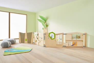 In einem hellen Raum mit hellgrünen Wänden stehen ein hölzerner Laufstall mit interaktiven Paneelen, eine baumförmige Wanddekoration, ein grüner und gelber Teppich und zwei graue Bodenkissen in der Nähe eines großen Fensters.
