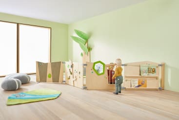 Ein Kleinkind steht in einem lichtdurchfluteten Raum neben einem hölzernen Laufstall mit interaktiven Elementen. Der Raum hat grüne Wände, eine Pflanzendekoration, Bodenkissen, ein Fenster und einen grünen Teppich auf Holzboden.
