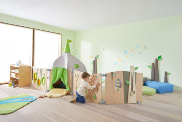 Ein kleines Kind spielt mit einem modularen Holzzaun in einem hellen, minimalistischen Spielzimmer. Es gibt ein Spielzelt in Form eines kleinen Hauses, weiche Sitzgelegenheiten und Wanddekorationen, die an Bäume und Blätter erinnern.