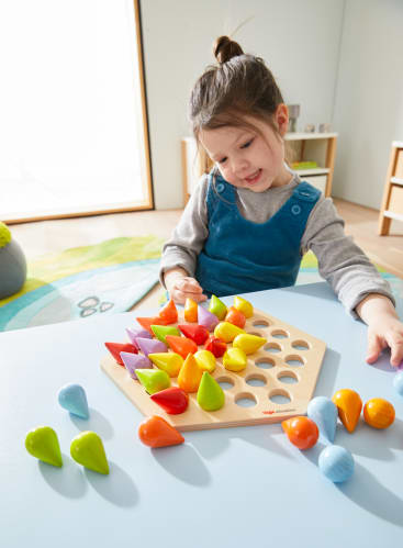 Ein junges Mädchen sitzt an einem Tisch und lächelt, während es in einem hellen, modernen Raum mit einem hölzernen Steckspielzeug mit bunten, kegelförmigen Teilen spielt.