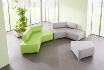 Ein modernes, modulares Sektionssofa mit grünen und hellgrauen Segmenten steht in der Ecke eines hellen Raums mit großen Fenstern, grauem Fußboden und einer hohen Topfpflanze. In der Nähe steht ein passender grauer Ottoman.