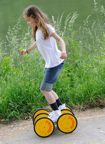 Ein Mädchen, das ein weißes Hemd, Jeansshorts und weiße Schuhe trägt, balanciert auf vier gelben, kreisförmigen Trittvorrichtungen mit der Aufschrift Pedalo auf einem Weg neben hohem Gras und Wasser.