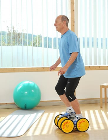 Ein älterer Mann in sportlicher Kleidung balanciert auf einem gelben Pedalo in einem hellen Raum mit großen Fenstern, einem grünen Gymnastikball, einer Matte und einem Holzstuhl im Hintergrund.