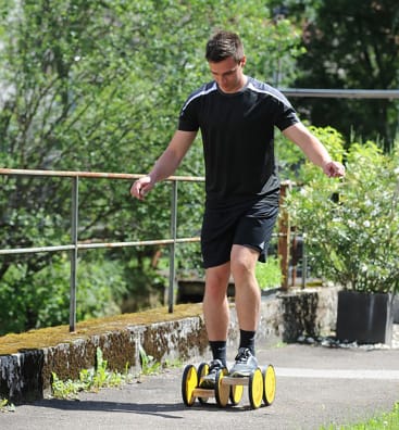 Ein Mann in schwarzer Sportkleidung balanciert auf einem Satz gelber und schwarzer Rollen, während er im Freien auf einem gepflasterten Weg mit viel Grün im Hintergrund spazieren geht.