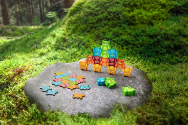 Bunte eulenförmige Spielzeugteile sind auf einem flachen Felsen gestapelt, der von grünem Moos in einem Wald umgeben ist. Weitere Teile, darunter Puzzleformen und Eulen, sind auf dem Felsen verstreut.