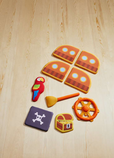Ein Set aus weichem Plüschspielzeug mit Piratenthematik ist auf einem hellen Holzboden angeordnet. Dazu gehören ein Papagei, eine Schatztruhe, ein Schiffsrad, ein Fernrohr, eine Piratenflagge und vier Teile, die an ein Schiff erinnern.