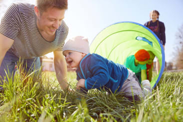 Ein Mann und ein Baby krabbeln im Gras neben einem grünen Spieltunnel. Ein anderes Kind krabbelt in den Tunnel hinein, während ein Erwachsener im Hintergrund steht. Die Szene spielt sich an einem sonnigen Tag im Freien ab.