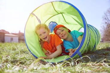 Zwei Kinder lächeln, während sie in einem grünen Spieltunnel auf einer Wiese im Freien liegen. Beide tragen bunte Hemden, und die Sonne scheint, so dass eine helle, fröhliche Szene entsteht.