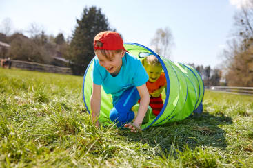 Zwei Kinder krabbeln durch einen grünen Spieltunnel auf einer Wiese. Das Kind vorne trägt eine rote Mütze und ein blaues Hemd, während das Kind hinten ein gelbes Hemd trägt. Im Hintergrund sind Bäume und ein Zaun zu sehen.