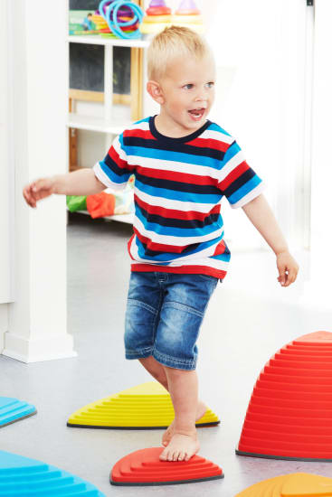 Ein kleiner Junge mit blondem Haar, der ein gestreiftes T-Shirt und Jeans-Shorts trägt, steht barfuß auf bunten Plastiktrittsteinen in einem hellen Indoor-Spielbereich.