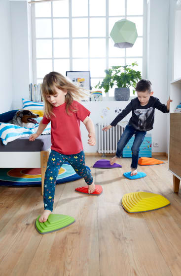 Zwei Kinder spielen in einem Zimmer und treten barfuß auf bunte Plastiktrittsteine, die auf dem Holzboden liegen. Das Zimmer hat ein Bett, eine Topfpflanze, Bücher und ein großes Fenster, das natürliches Licht hereinlässt.