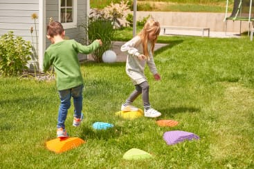 Zwei Kinder spielen draußen auf einer Wiese und balancieren auf bunten Plastiktrittsteinen, die in einer Reihe neben einem hellgrauen Haus angeordnet sind. Der Junge trägt Jeans und ein grünes Hemd, das Mädchen Leggings und ein graues Kleid.