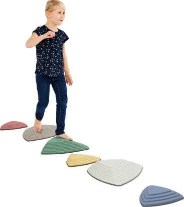 Ein kleines Kind läuft barfuß auf bunten, dreieckigen Trittsteinen, die in einer Reihe auf weißem Hintergrund angeordnet sind. Das Kind trägt ein marineblaues Hemd mit weißen Punkten und eine dunkle Jeans.