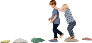 Zwei Jungen balancieren auf erhöhten, bunten Trittsteinen, die in einer Reihe angeordnet sind. Der vordere Junge geht voraus, während sich der hintere Junge an den Schultern abstützt. Beide sind barfuß und tragen Freizeitkleidung.