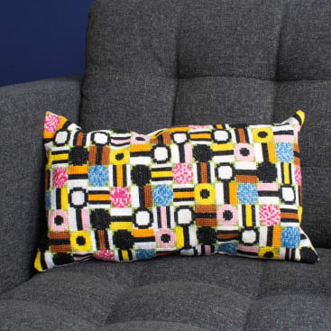 Ein rechteckiges Kissen mit einem bunten geometrischen Muster aus Kreisen und Rechtecken in Schwarz, Weiß, Gelb, Rosa und Blau liegt auf einer dunkelgrau gepolsterten Couch.