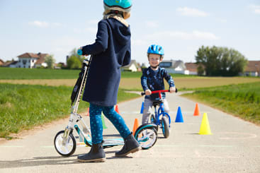 Zwei Kinder mit Helmen sind auf einem gepflasterten Weg im Freien unterwegs. Ein Kind mit einem Motorroller steht im Vordergrund, während ein anderes Kind im Hintergrund ein kleines Fahrrad fährt. Auf dem Weg sind bunte Kegel aufgestellt.