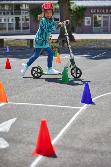 Ein Kind mit einem roten Helm fährt auf einem grünen Roller durch einen mit bunten Verkehrskegeln markierten Parcours auf einer Asphaltfläche. Im Hintergrund sind ein Baum, ein Gebäude und Bänke zu sehen.