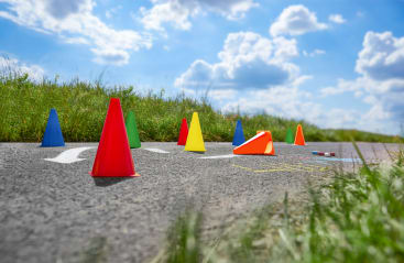 Mehrere bunte Verkehrskegel, darunter rote, gelbe, blaue, grüne und orangefarbene, sind auf einem asphaltierten Weg im Freien mit Gras auf beiden Seiten unter einem teilweise bewölkten Himmel angeordnet und umgestoßen.