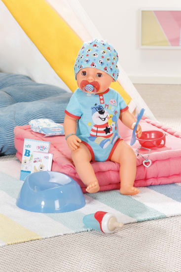 Eine Babypuppe mit blauer Mütze und Strampler sitzt auf rosa Handtüchern und hält einen Schnuller und einen Löffel in der Hand. Um sie herum stehen ein blaues Töpfchen, eine Babyflasche, Essensdosen und ein Spielzeugstethoskop in einer Spielzimmerumgebung.