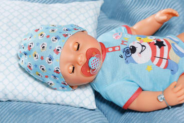 Eine Babypuppe mit blauer Mütze und passendem T-Shirt mit Cartoon-Tieren liegt auf einem blauen Kissen und einer blauen Decke und hält einen roten Schnuller im Mund.