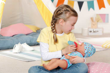 Ein junges Mädchen mit geflochtenen Haaren und einem gelben Hemd sitzt im Schneidersitz auf dem Boden und füttert eine Babypuppe mit einer Spielzeugflasche in einem hellen, farbenfrohen Spielzimmer mit Kissen und Wimpeln.