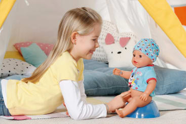 Ein junges Mädchen liegt in einem Haus auf dem Bauch, lächelt und spielt mit einer Babypuppe in einem blauen Outfit mit Hut. Die Puppe sitzt auf einem Spielzeug-Töpfchen. Im Hintergrund sind Kissen und ein Zelt zu sehen.