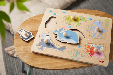 Auf einem kleinen runden Holztisch liegt ein Holzpuzzle mit Meerestieren, darunter ein Pinguin, ein Wal, eine Robbe, eine Schildkröte, ein Oktopus und eine Krabbe. Ein Teil, der Pinguin, ist herausgenommen und liegt neben dem Brett.