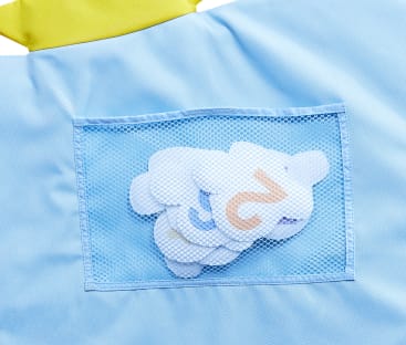 Eine Netztasche auf hellblauem Stoff enthält ein kleines weißes Plüschtier mit gelben Markierungen. Oben auf dem Bild ist ein Teil eines gelben Kragens oder einer gelben Verzierung zu sehen.