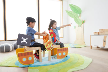 Zwei Kinder sitzen auf als Piratenschiffe dekorierten Stühlen in einem hell erleuchteten Spielzimmer. Ein Kind trägt einen schwarzen Piratenhut, das andere zeigt nach vorne und hält einen Papagei aus Stoff. Es gibt grüne Teppiche und Wanddekorationen in Form von Bäumen.